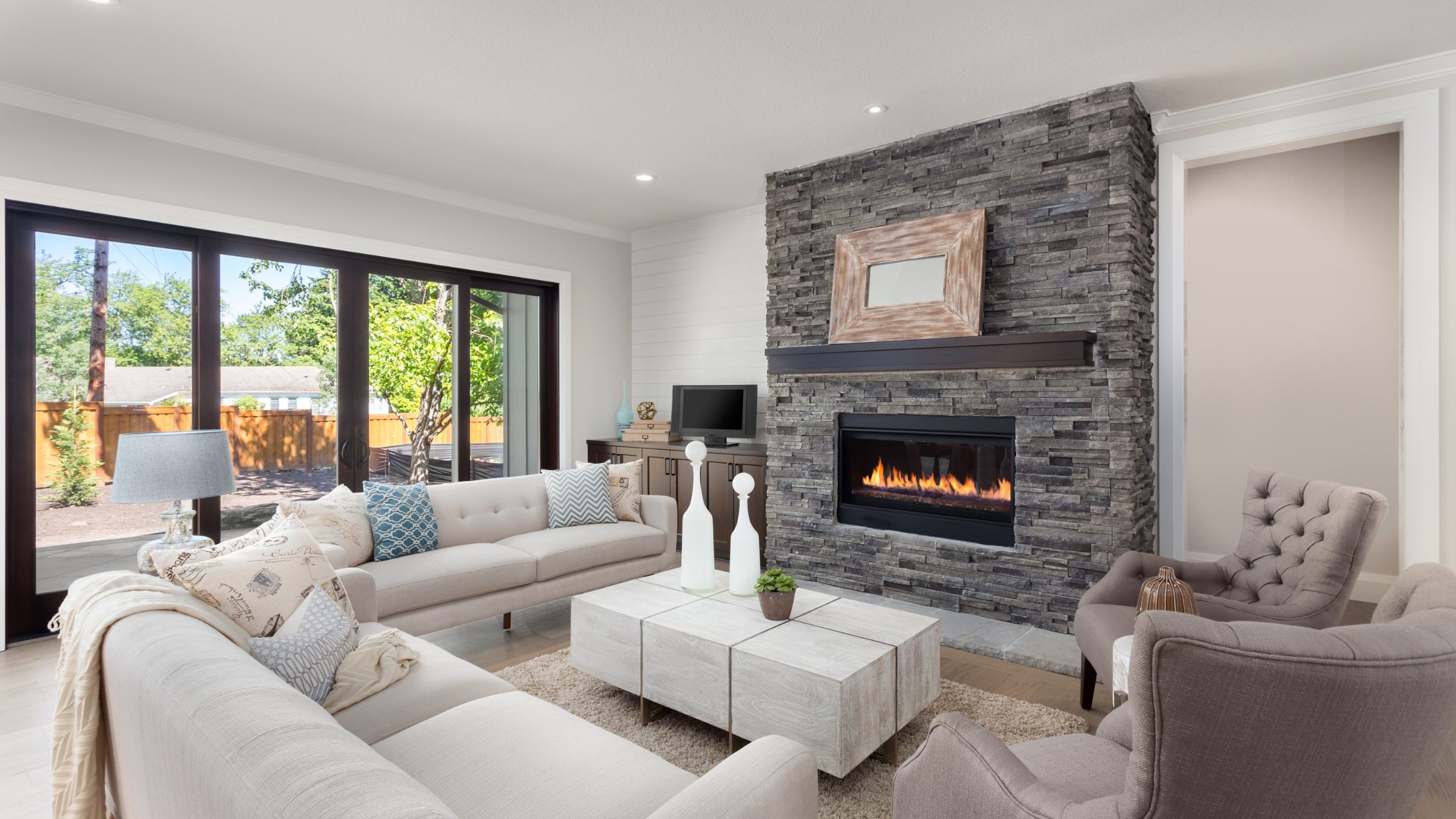 Stock photo of a living room. White couches, a lit fireplace with grey brick, a white coffee table, and a small grey seat for one on the right. The room is bright.