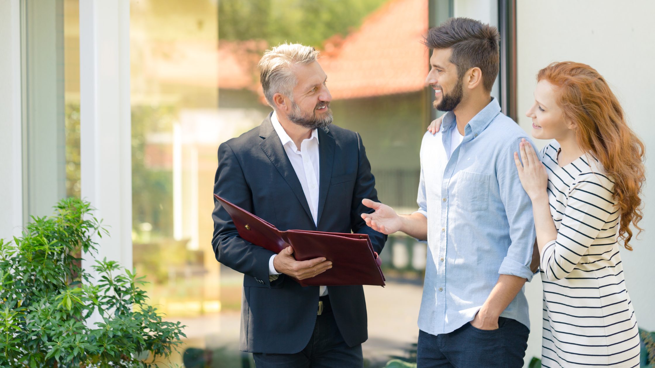 Stock image of a young couple (right) speaking with a realtor (left). The couple is a man and woman, man is wearing a light blue shirt on the left of the woman who is wearing a black and white striped long-sleeved shirt with her arm resting on the man's shoulder. The Realtor is on the left of the couple speaking to them while holding open a maroon folder.