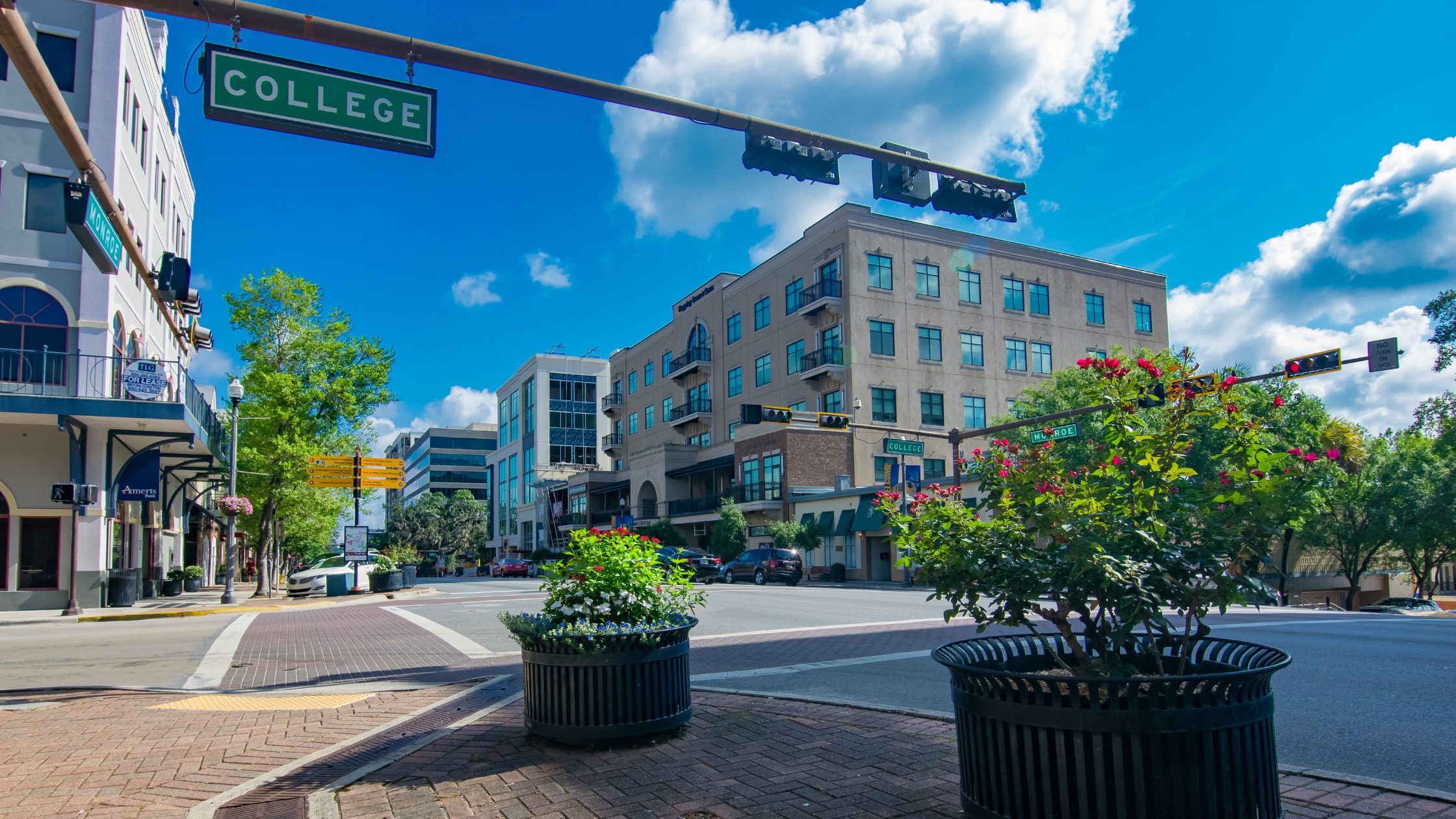 Stock image of Downtown Tallahassee, Florida, on College Street. The photo is taken from a corner sidewalk looking towards a building and a road with traffic lights in between. The sky is blue and cloudy in the background. There are lots of trees and potted plants around.