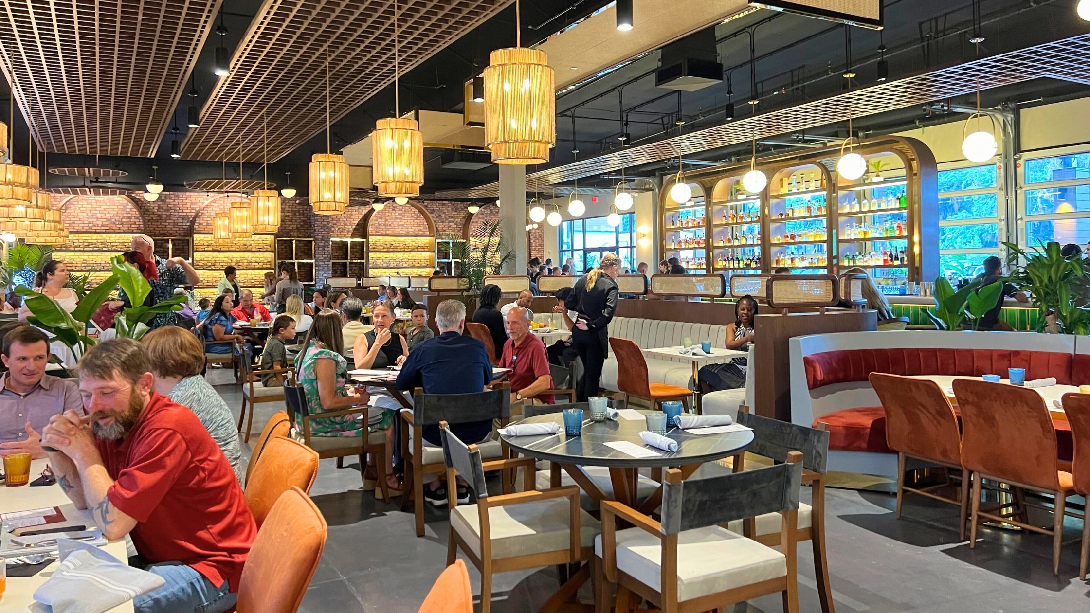 Photo taken at The Monroe restaurant in Tallahassee, Florida. The photo was taken inside and shows the dining room with people sitting, talking, and eating at their tables. It is during the day because you can see the daylight through the windows in the back of the room.